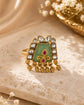 A gold ring with a green enamel inlay, a central red jewel, and clear stones, styled on a textured beige stone with cream silk and dried white flowers.