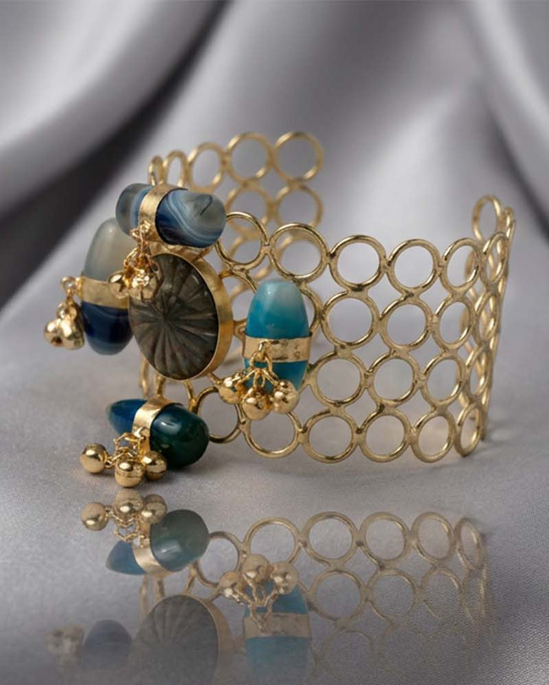 A gold-tone open-link cuff bracelet with turquoise and dark gray cabochon stones, displayed on draped silver satin fabric.